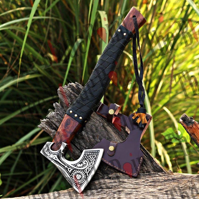 May include: A handcrafted axe with a wooden handle and a black leather-wrapped grip. The axe head is silver with intricate engraved designs. A leather sheath and a black lanyard are attached. The axe is displayed on a piece of wood.