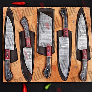 Pu&ograve; includere: Set di cinque coltelli da cucina con lame in acciaio damascato e manici in legno. I coltelli sono esposti in foderi di cuoio su uno sfondo marrone.