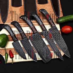 Pu&ograve; includere: Set di cinque coltelli da cucina neri con manici in legno e lama testurizzata. I coltelli sono disposti su un tagliere di legno con un cetriolo, un pomodoro e un peperoncino.