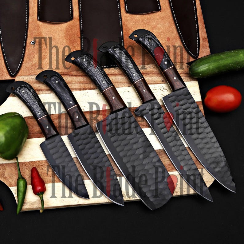 Pu&ograve; includere: Set di cinque coltelli da cucina neri con manici in legno e lama testurizzata. I coltelli sono disposti su un tagliere di legno con un cetriolo, un pomodoro e un peperoncino.