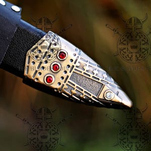 May include: A black leather sheath tip featuring an ornate brass-toned metal fitting with three embedded red circular stones and one clear stone at the point. The metal surface displays intricate etched patterns and geometric detailing.