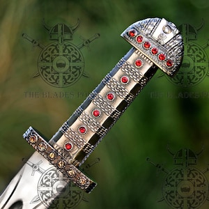 May include: A metallic sword hilt featuring a textured, grid-like pattern adorned with multiple small, circular red gemstones embedded along the grip and the pommel. The crossguard is decorated with a row of smaller, amber-colored stones.