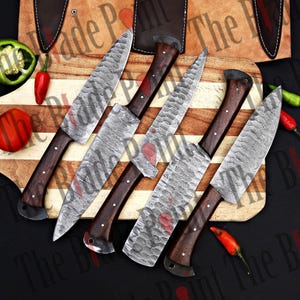 Pu&ograve; includere: Cinque coltelli da cucina in acciaio Damasco con manici in legno su un tagliere di legno. I coltelli hanno una finitura martellata e sono disposti a ventaglio.