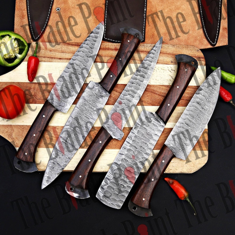 Pu&ograve; includere: Cinque coltelli da cucina in acciaio Damasco con manici in legno su un tagliere di legno. I coltelli hanno una finitura martellata e sono disposti a ventaglio.