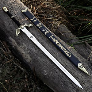 May include: A silver-toned metal sword with a black-wrapped handle, a gold-colored crossguard shaped like wings, and a triforce symbol on the blade. It is accompanied by a black scabbard featuring gold-colored decorative engravings and symbols.