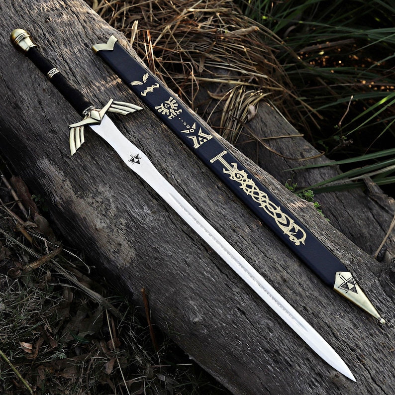 May include: A silver-toned metal sword with a black-wrapped handle, a gold-colored crossguard shaped like wings, and a triforce symbol on the blade. It is accompanied by a black scabbard featuring gold-colored decorative engravings and symbols.
