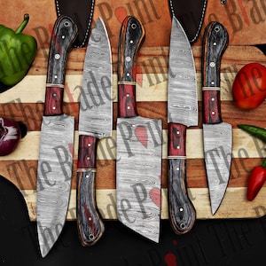 Pu&ograve; includere: Set di cinque coltelli da cucina con lame in acciaio Damasco e manici in legno. Le lame hanno un motivo ondulato e i manici sono neri e rossi.