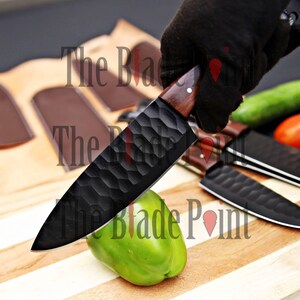 Pu&ograve; includere: Un coltello da chef nero con manico in legno e lama testurizzata viene utilizzato per tagliare un peperone verde su un tagliere di legno. Il coltello &egrave; affilato e ha un design distintivo.
