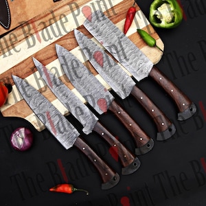 Pu&ograve; includere: Set di cinque coltelli da cucina in acciaio damascato con manici in legno. I coltelli sono disposti su un tagliere di legno con peperoni rossi e verdi.