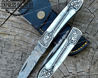 May include: Two ornate pocket knives with intricate floral designs on the blades and handles. One knife has a Damascus steel blade, while the other has a polished steel blade. Both knives are in a leather sheath.