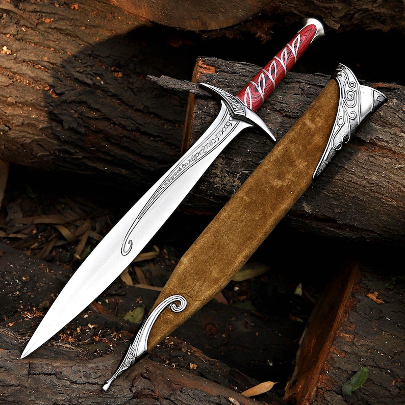 May include: A silver-toned metal sword with a pointed blade featuring etched script, a red and black patterned handle, and a silver pommel. The sword is accompanied by a tan suede-textured scabbard with ornate silver metal accents.