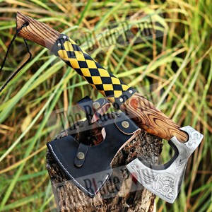 May include: A handcrafted axe with a wooden handle and a black and yellow checkered grip. The axe head is silver with an engraved design. A black leather sheath is included. The axe is displayed on a wooden stump.