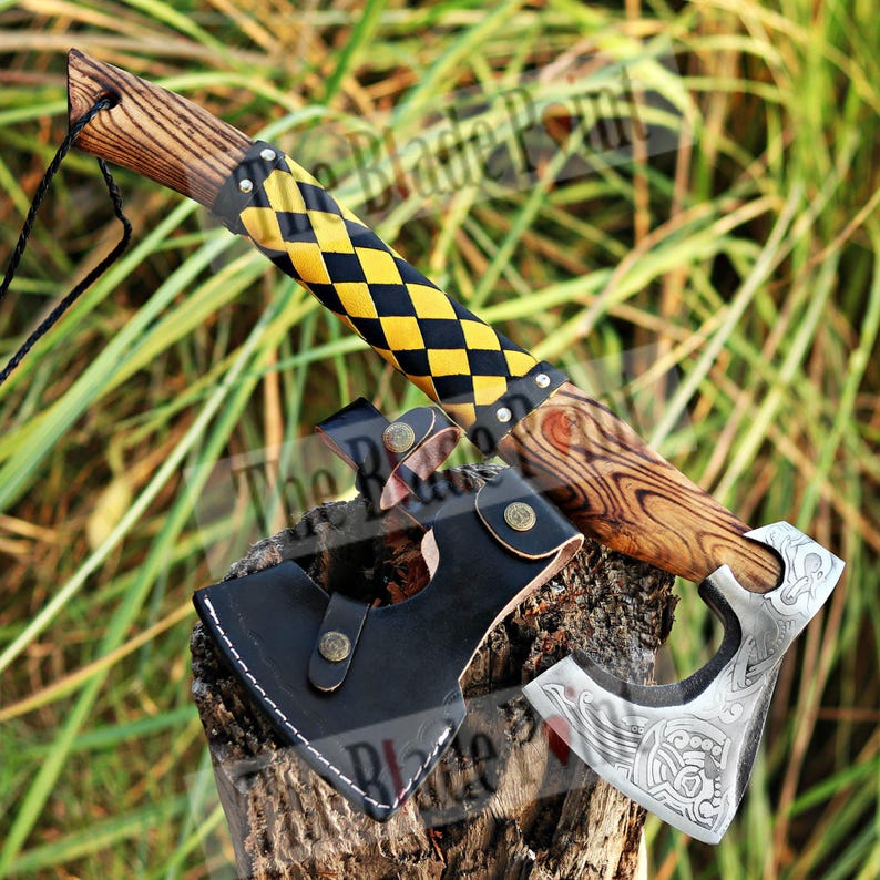 May include: A handcrafted axe with a wooden handle and a black and yellow checkered grip. The axe head is silver with an engraved design. A black leather sheath is included. The axe is displayed on a wooden stump.