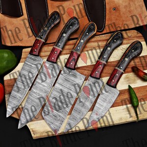 Pu&ograve; includere: Set di cinque coltelli da cucina con lame in acciaio damascato e manici in legno. Le lame hanno un motivo ondulato e i manici sono neri, rossi e marroni.