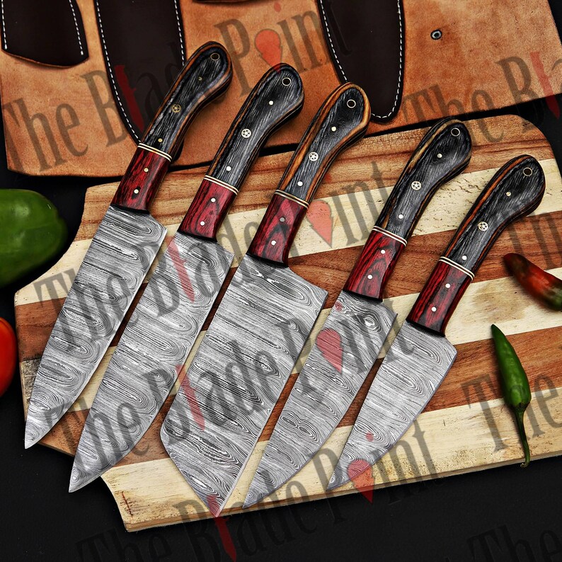 Pu&ograve; includere: Set di cinque coltelli da cucina con lame in acciaio damascato e manici in legno. Le lame hanno un motivo ondulato e i manici sono neri, rossi e marroni.