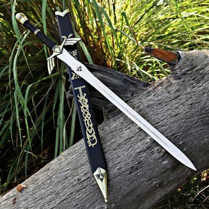 May include: A silver-toned metal sword with a black handle and a gold-colored crossguard shaped like wings. The blade features a small triangular emblem. It is paired with a black scabbard decorated with gold-colored intricate patterns.