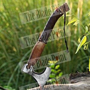 May include: A Viking axe with a wooden handle and a silver-colored blade. The handle is wrapped in dark brown leather. The blade features intricate engraved designs. A black cord is attached to the handle. The axe is displayed outdoors.