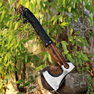 May include: A handcrafted axe with a wooden handle and a black leather-wrapped grip. The axe head is silver with an engraved design. The axe is displayed on a wooden surface against a backdrop of green foliage.