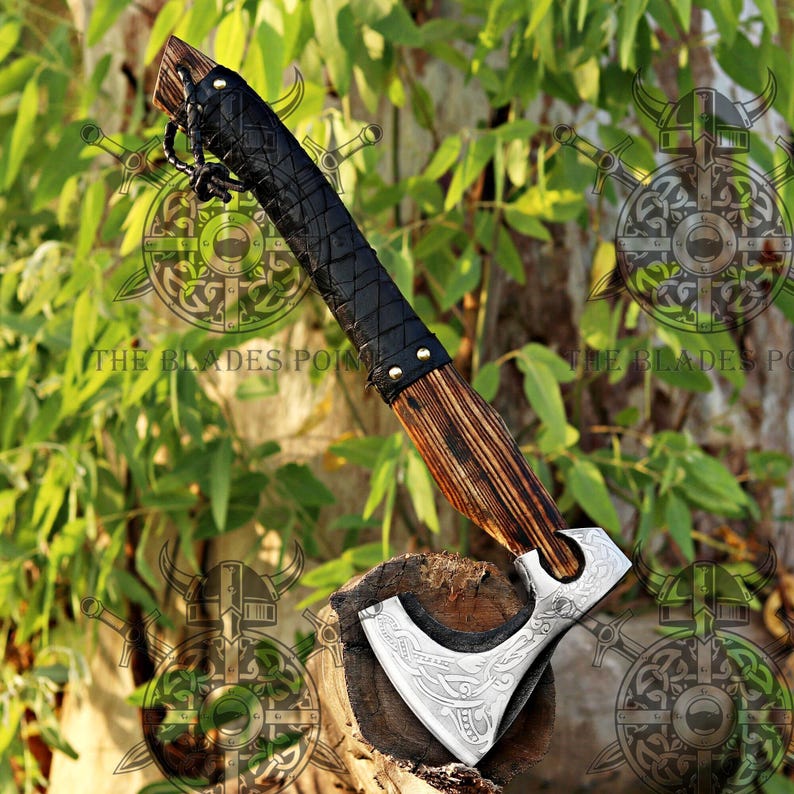 May include: A handcrafted axe with a wooden handle and a black leather-wrapped grip. The axe head is silver with an engraved design. The axe is displayed on a wooden surface against a backdrop of green foliage.