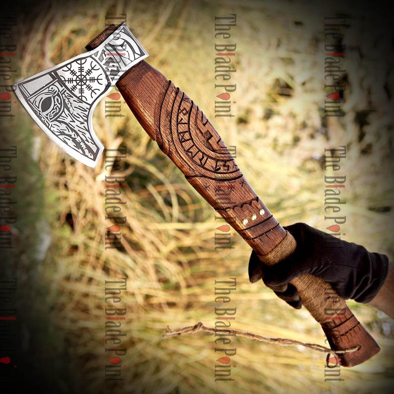 May include: A handcrafted axe with a wooden handle and a silver-colored blade. The blade features etched Viking-inspired designs, including runes and a compass symbol. The handle is intricately carved with patterns and wrapped with twine.