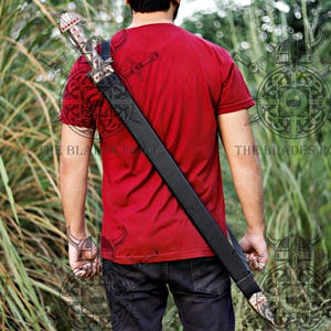 May include: A long sword in a black leather scabbard with ornate metal fittings featuring red accents, worn diagonally across the back. The sword hilt has intricate metalwork and red inlays, and the scabbard has a matching metal tip.