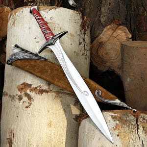 May include: A silver-toned metal sword with a pointed blade featuring etched script, a crossguard, and a red handle with silver accents. The sword is accompanied by a brown suede scabbard with ornate silver-toned metal fittings at the ends.