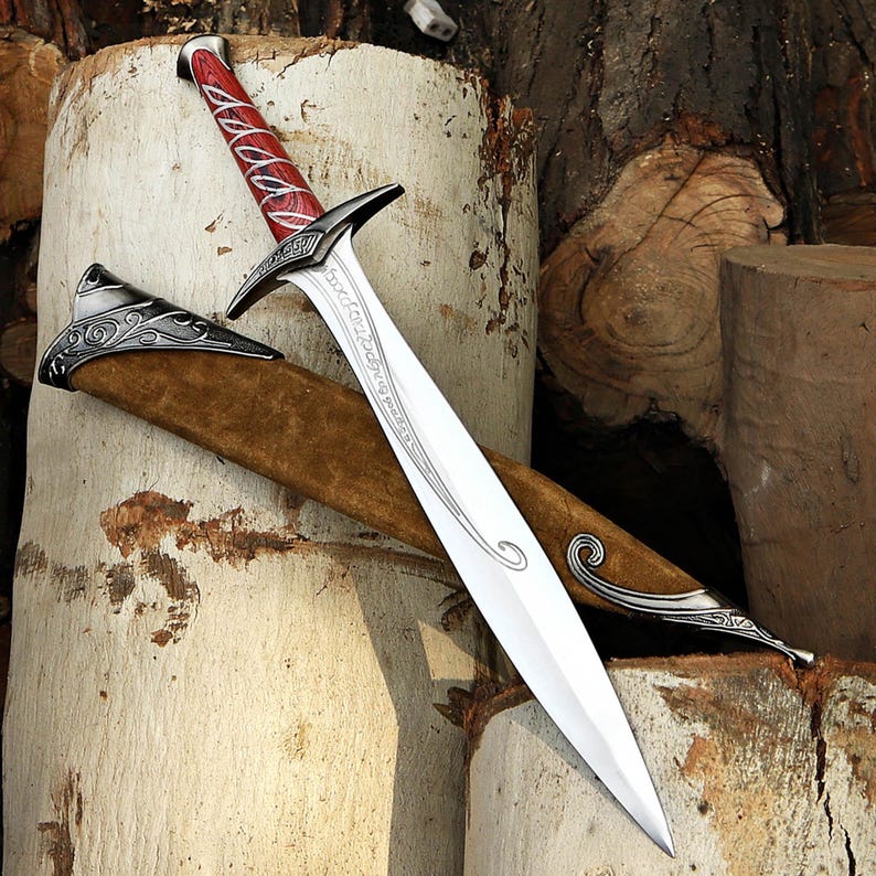 May include: A silver-toned metal sword with a pointed blade featuring etched script, a crossguard, and a red handle with silver accents. The sword is accompanied by a brown suede scabbard with ornate silver-toned metal fittings at the ends.