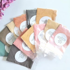 May include: A collection of small, clear plastic pouches filled with various colored powders, arranged in a fan shape. Each pouch features a circular white label with printed text. The colors include shades of pink, yellow, gray, green, and tan.