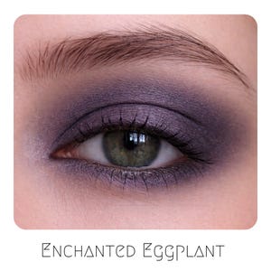 May include: An eye with dark purple eyeshadow applied in a smoky style across the eyelid and crease, featuring a shimmer finish. The text ENCHANTED EGGPLANT is printed in a thin, black, sans-serif font at the bottom of the image frame.