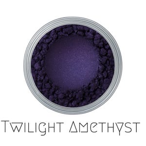 May include: A round container filled with loose, deep purple powder pigment featuring a shimmering, metallic finish. The powder has a textured, granular appearance around the edges and a smooth center. The text TWILIGHT AMETHYST is below.