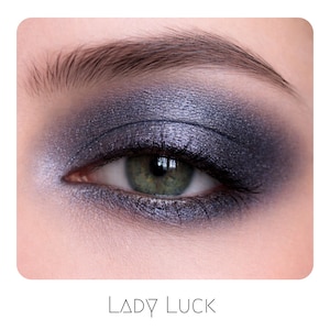 May include: An eye with a green iris and dark purple metallic eyeshadow applied in a smoky style across the eyelid and lower lash line. The text LADY LUCK is printed in black below the eye. The makeup features a shimmering finish with deep tones.