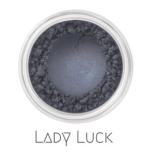 May include: A round container filled with loose, dark grey, shimmering cosmetic powder. The center of the powder is pressed into a smooth, circular shape, while the edges remain textured and loose. The text LADY LUCK is printed below.