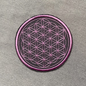 May include: A round, black embroidered patch with a pink flower of life design. The patch has a pink border.