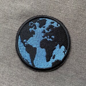 Könnte beinhalten: Ein blauer und schwarzer gestickter Aufnäher, der eine Weltkarte darstellt. Der Aufnäher ist rund und hat einen schwarzen Rand.