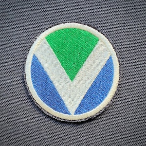 Può includere: Una toppa ricamata bianca, verde e blu a forma di cerchio con una V bianca al centro. La V è circondata da un triangolo verde e una forma a mezzaluna blu.