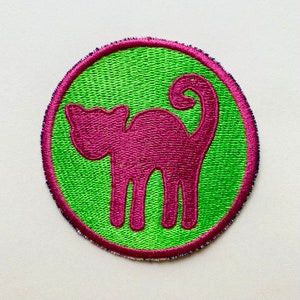 Könnte beinhalten: Ein runder grüner Patch mit einer rosa gestickten Katzensilhouette. Die Katze schaut nach links, der Schwanz ist eingerollt.