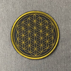 Puede incluir: Un parche bordado en negro y dorado con un diseño de flor de la vida. El parche es circular y tiene un contorno dorado.