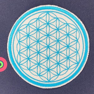 Puede incluir: Un parche de tela blanca con un diseño de flor de la vida bordado en azul. El parche tiene un borde azul y es circular.