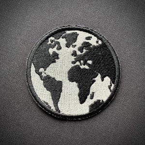 Pode incluir: Um remendo bordado preto e branco da Terra com um contorno branco. O remendo é circular e tem uma borda preta.
