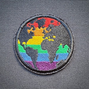 Könnte beinhalten: Ein runder gestickter Aufnäher mit einer schwarzen Umrandung der Welt in Regenbogenfarben. Der Aufnäher ist auf einen dunkelgrauen Stoff genäht.