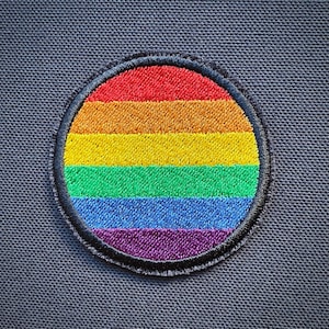 Può includere: Una toppa circolare arcobaleno pride con un bordo nero. La toppa è realizzata in tessuto e ha sei strisce di rosso, arancione, giallo, verde, blu e viola.