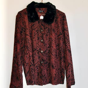 Può includere: Giacca fantasia rossa e nera con colletto in finta pelliccia nera. La giacca ha una chiusura con bottoni sul davanti e maniche lunghe. Il motivo è a spina di pesce. La giacca è appesa a una gruccia color oro.