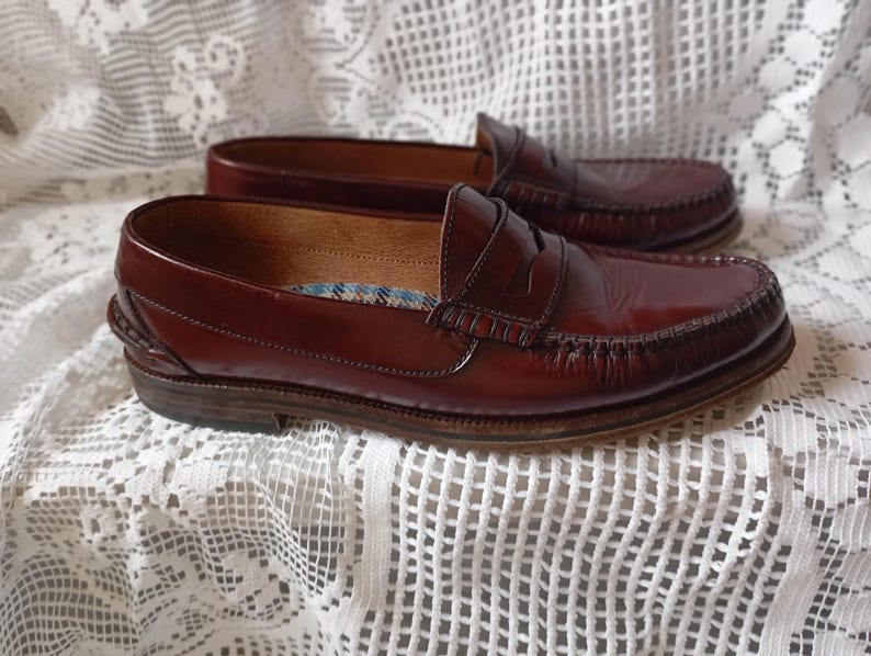 May include: A pair of burgundy leather penny loafers. The shoes feature a classic design with a rounded toe, a strap across the vamp, and a stitched sole. The leather has a glossy finish. The shoes appear to be in good condition.