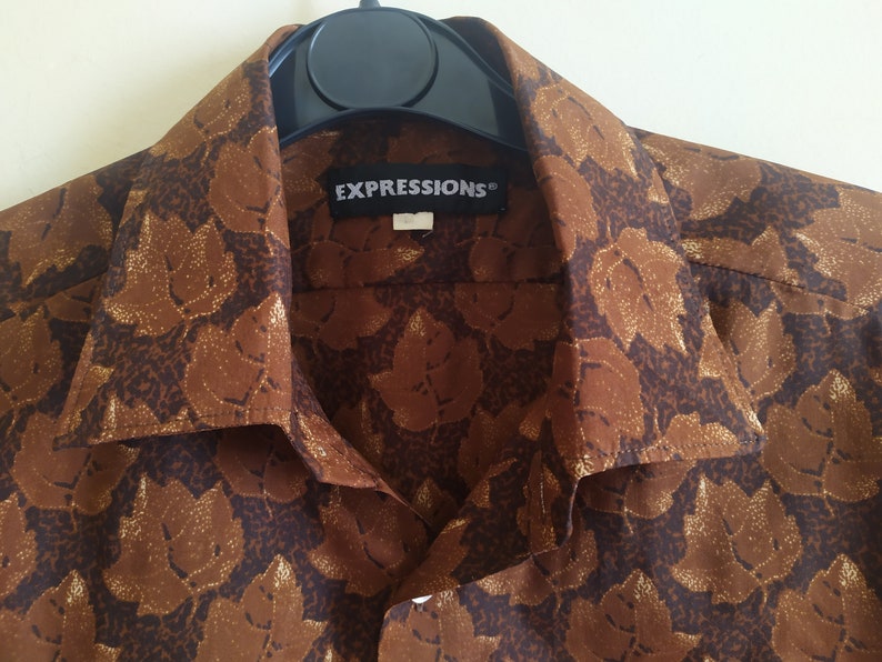 Puede incluir: Una camisa marr&oacute;n de botones con un estampado de hojas doradas. La camisa tiene cuello y una etiqueta que dice "EXPRESSIONS".