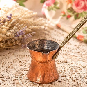 May include: A hammered copper coffee pot with a wide base, a narrow neck, and a flared rim. The pot features a long, slender, decorative brass handle attached with a single rivet. The interior of the pot has a metallic silver-toned lining.