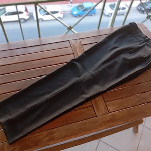 May include: A pair of solid black trousers featuring a structured waistband, a front button closure, and a clean, flat-front design. The fabric has a smooth, matte finish with visible pressed creases running down the centre of each leg.
