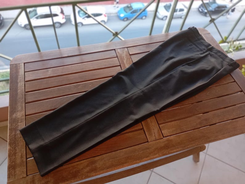 May include: A pair of solid black trousers featuring a structured waistband, a front button closure, and a clean, flat-front design. The fabric has a smooth, matte finish with visible pressed creases running down the centre of each leg.