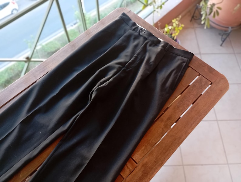 May include: A pair of solid black trousers featuring a structured waistband with a single button closure, a front zip fly, and a clean, straight-leg cut with pressed creases running down the centre of each leg, creating a formal appearance.