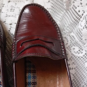 May include: Close-up of a pair of burgundy leather penny loafers. The shoes have a classic design with a stitched vamp and a decorative strap across the top. The interior features a plaid pattern.