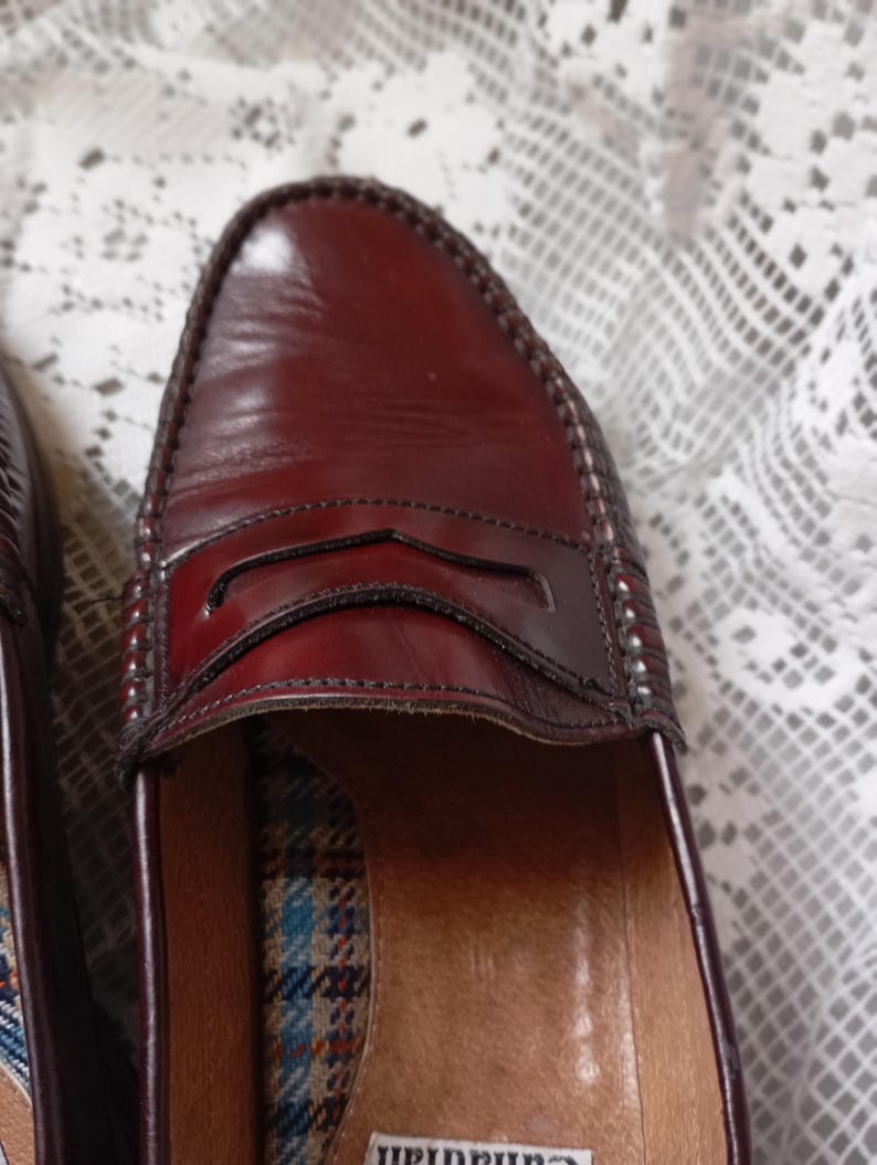 May include: Close-up of a pair of burgundy leather penny loafers. The shoes have a classic design with a stitched vamp and a decorative strap across the top. The interior features a plaid pattern.